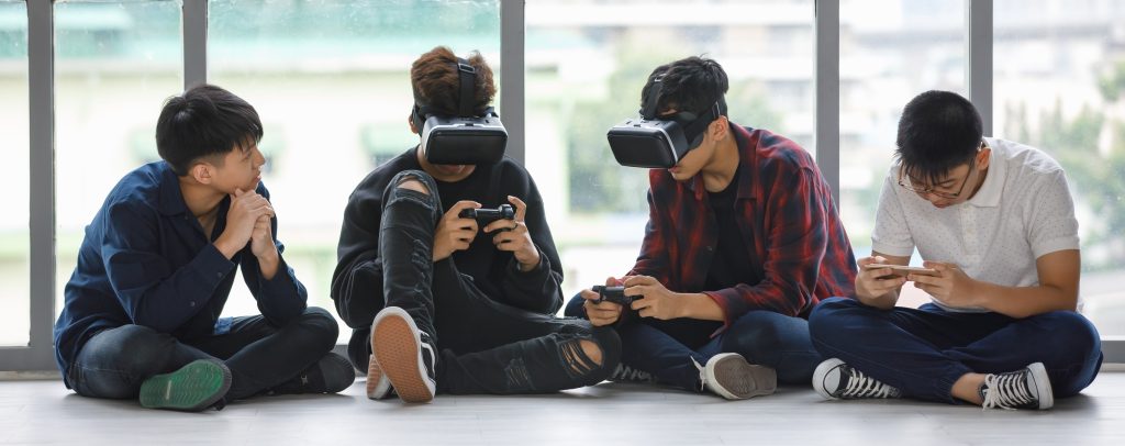 Group of four handsome young male teenagers with cute smiling sitting on floor together. Junior boys playing games with virtual reality headset, controllers, and smartphones. Concept of web 3 gaming and technology