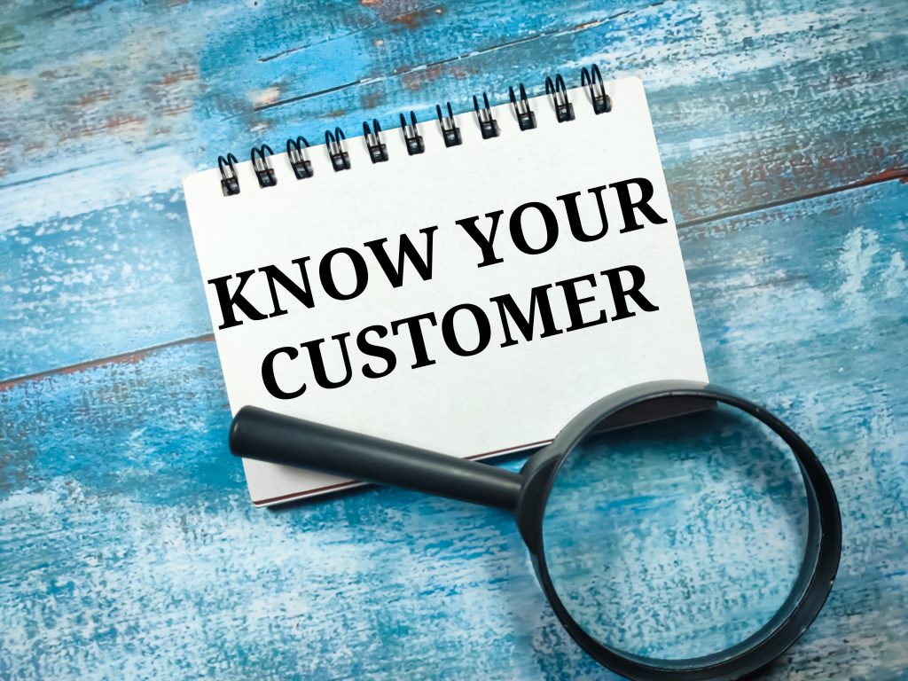 Text KNOW YOUR CUSTOMER written on notebook with magnifying glass on blue wooden background.