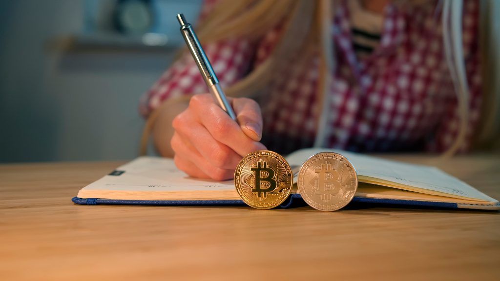 Crop view of female hands in checkered shirt holding shiny metal pen, and writing into daily planner with bitcoins placed on edges on foreground at night.Learning about the web3.