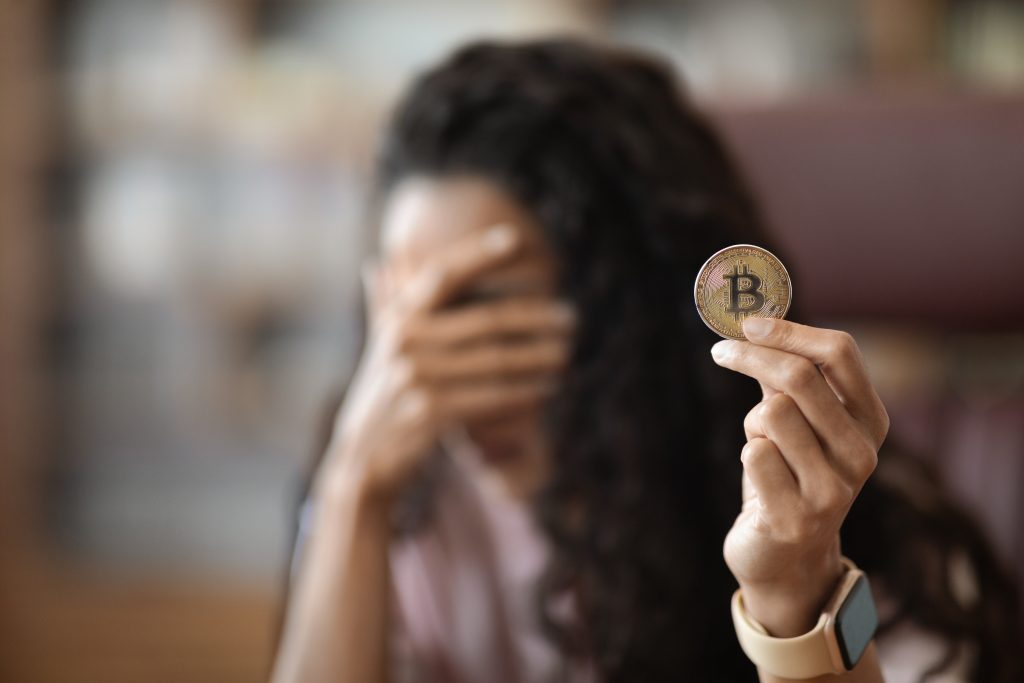 Young brunette woman holding virtual currency bitcoin at the office stressed and frustrated with hand covering her face, disappointed and depressed, blurred background, copy space