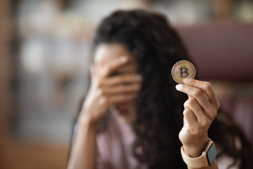 Mujer joven morena que sostiene bitcoin de moneda virtual en la oficina estresó y frustrada con la mano cubriendo su rostro, decepcionada y deprimida, antecedentes borrosos, espacio de copia