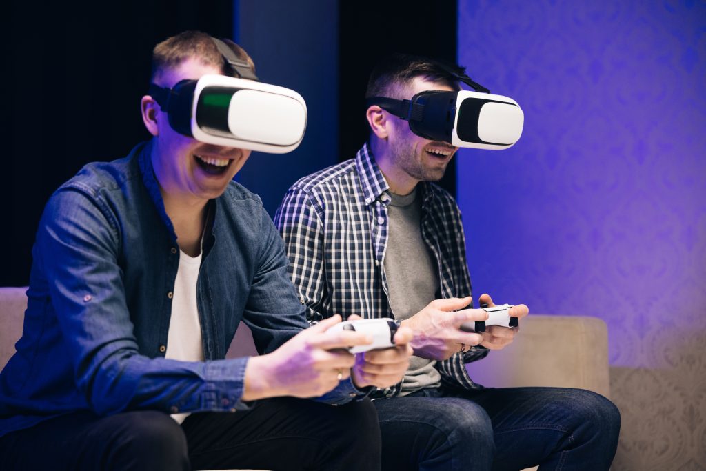 Excited funny young gamers wear virtual reality glasses holding controllers playing video game. Overjoyed men friends winning videogame having fun together on sofa celebrating victory at home.