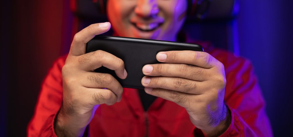 Jugar videojuegos en el teléfono inteligente. Un joven apuesto asiático sentado en una silla sosteniendo un celular en la sala de estar. Felicidad Streamer Hombre indio con auriculares jugando en línea en el cuarto oscuro.