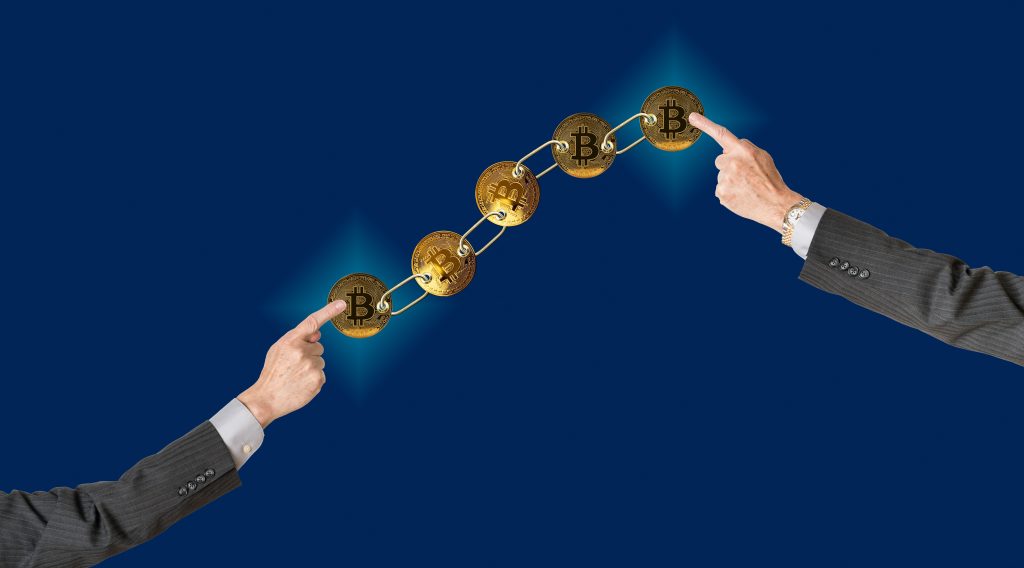 Conjunto de bitcoins dorados vinculados por cadena sobre fondo azul con dos brazos que apuntan a fines para ilustrar el concepto de blockchain para la gestión de la cadena de suministro